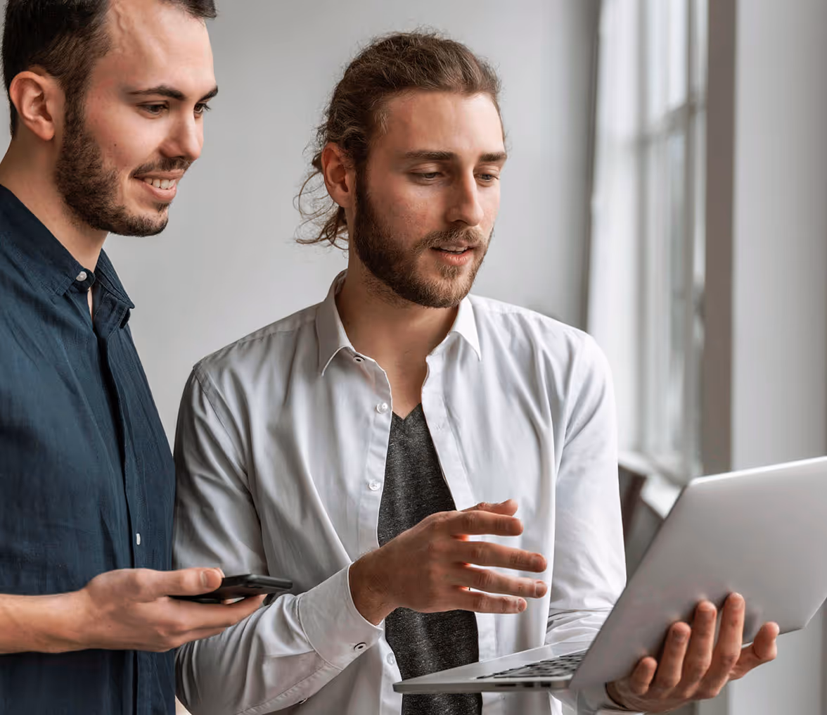 Two men looking at a laptop screen while one holds the laptop and the other holds a smartphone.  - QFS XLM